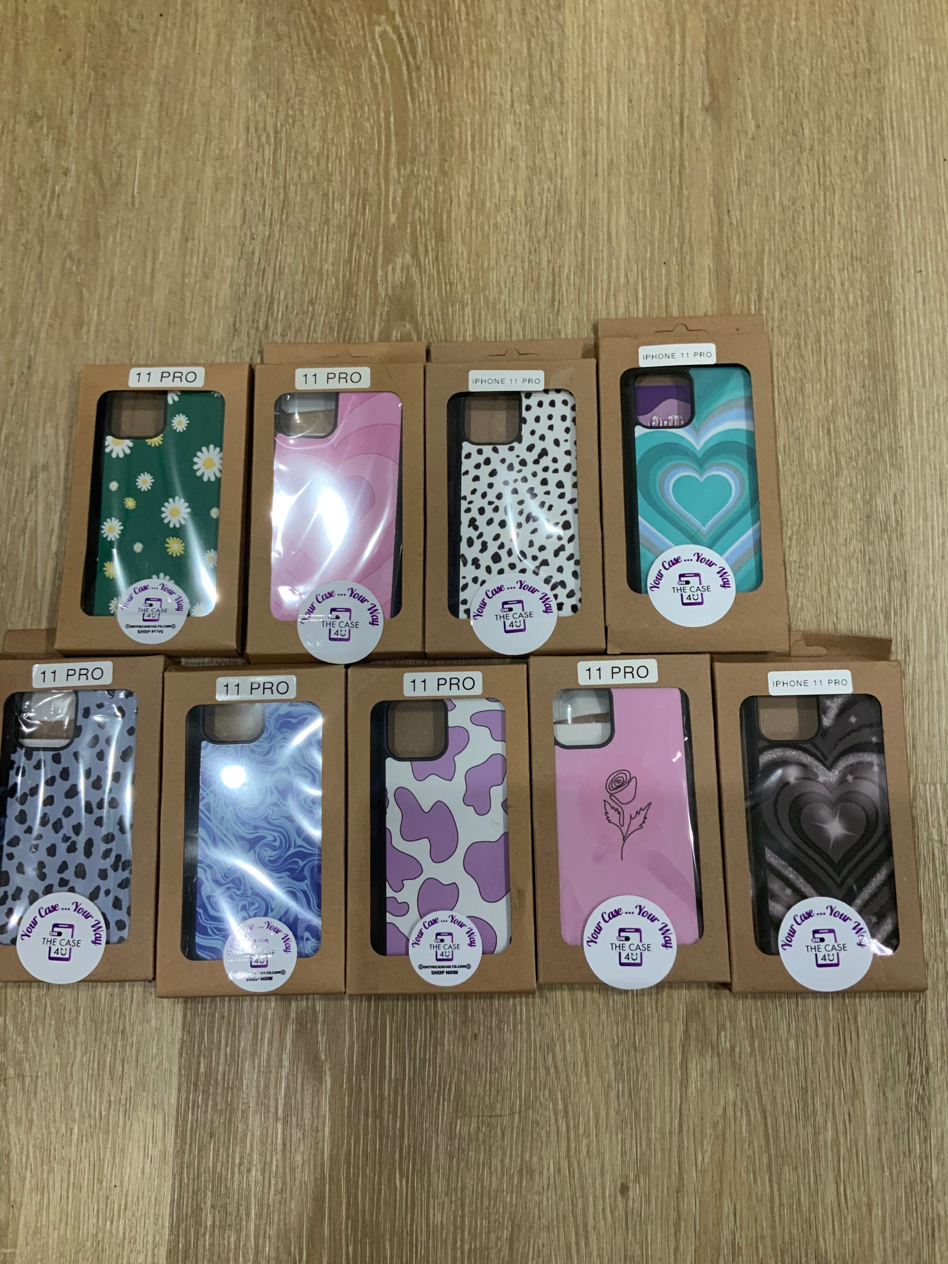 A collection of phone cases with various designs, packaged in cardboard boxes. Visible cases have patterns such as cow print, hearts, and floral designs.